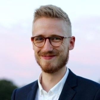 En mand med skæg iført briller og jakkesæt smiler til kameraet.