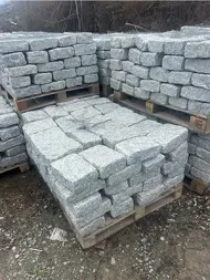 Three wooden pallets stacked with rectangular, light-gray granite pavers outdoors on a dirt ground.