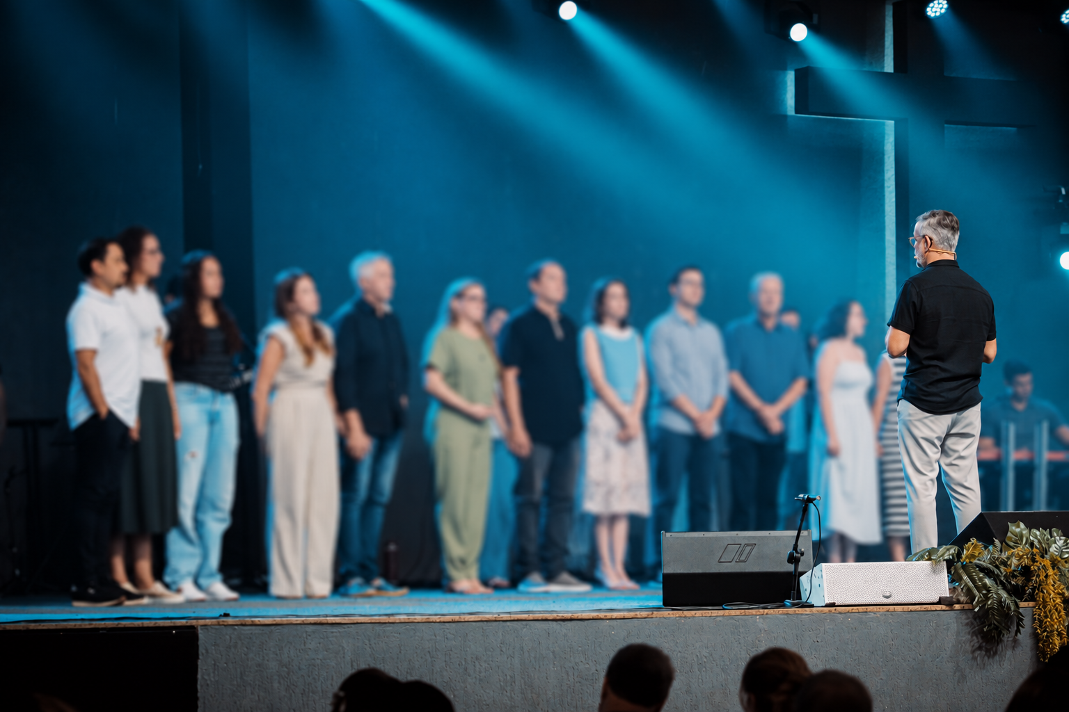Comunidade reunida no culto, com o pastor de costas e a cruz em foco; demais pessoas suavemente desfocadas