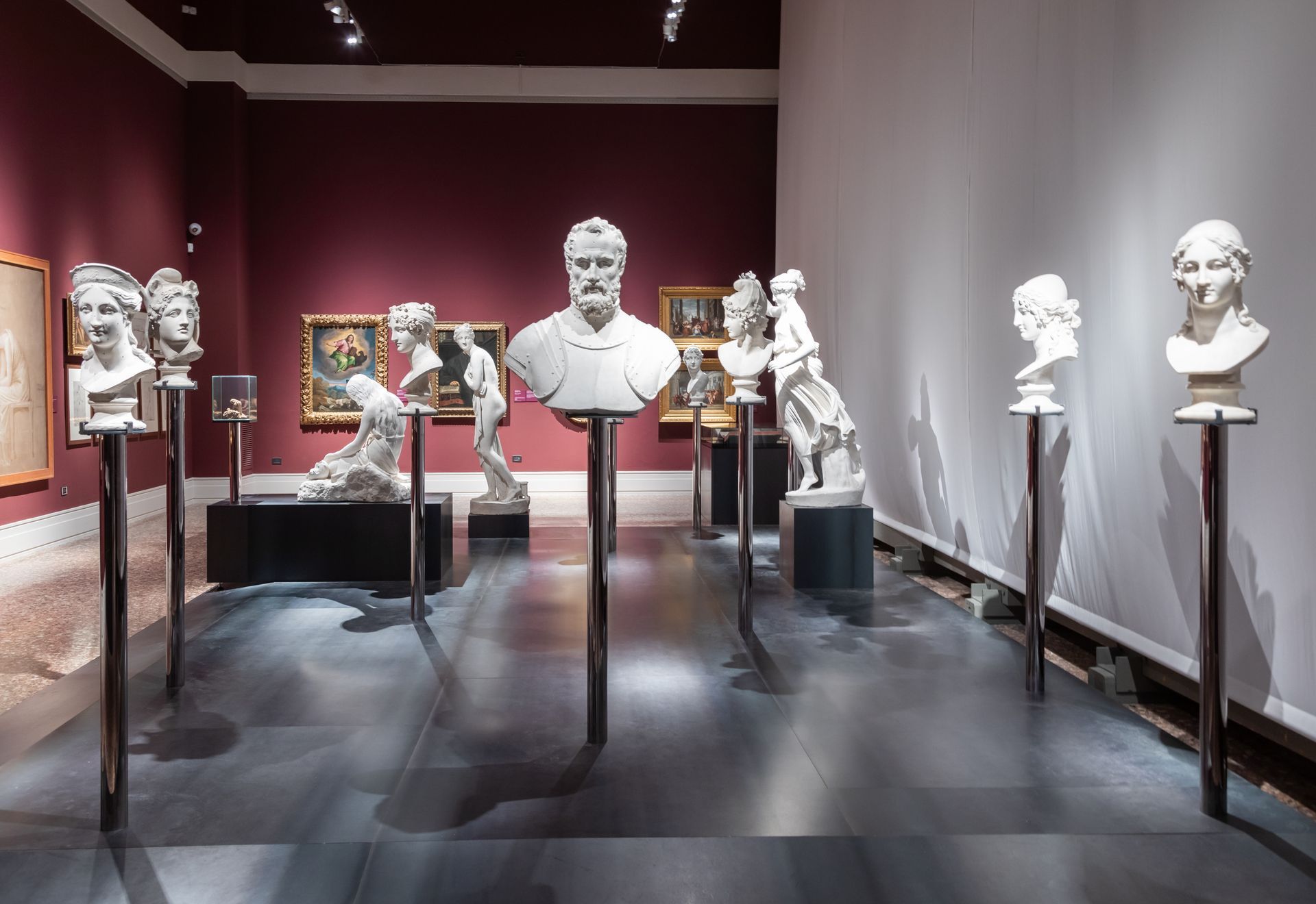 Bustos de mármore branco em altos postes de metal são exibidos em uma galeria com paredes e pinturas em vermelho escuro.