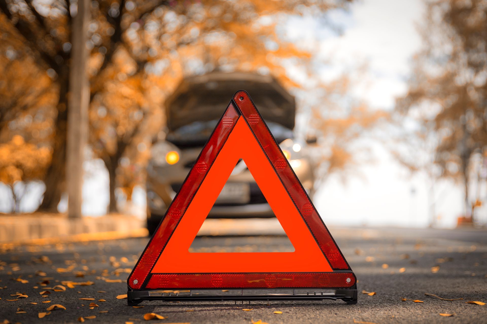 Triângulo de advertência vermelho está posicionado em uma estrada asfaltada em frente a um carro com o capô levantado.