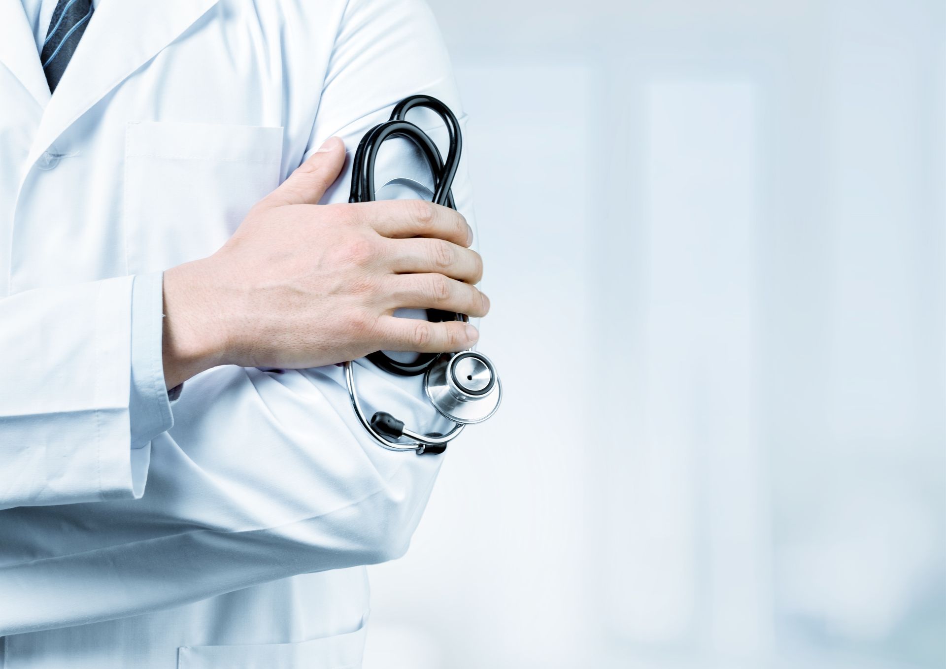 Um médico de jaleco branco segurando um estetoscópio com os braços cruzados, contra um fundo médico desfocado.