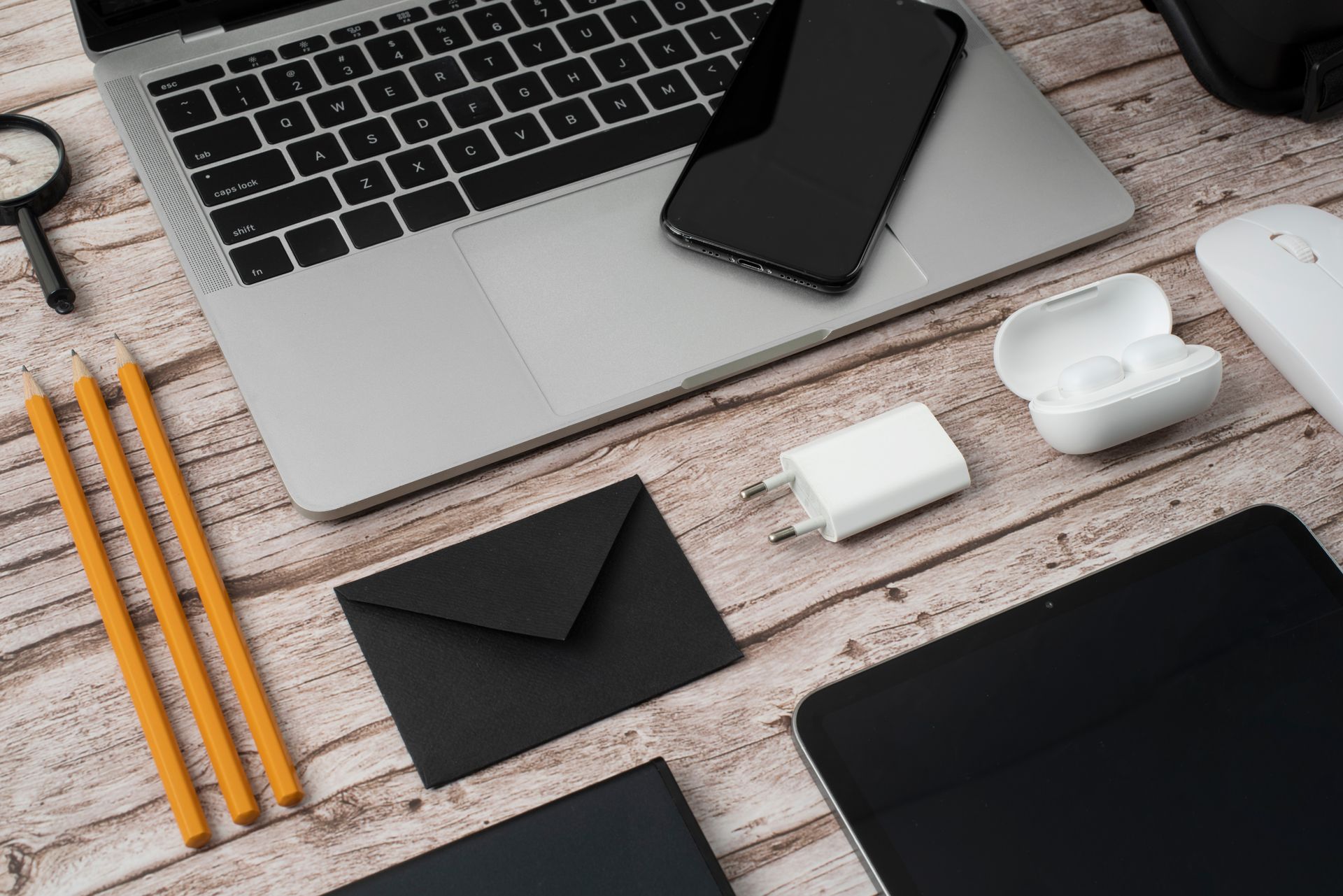 Uma composição de um laptop, smartphone, carregador, fones de ouvido, lápis, envelopes e tablets sobre uma mesa de madeira.