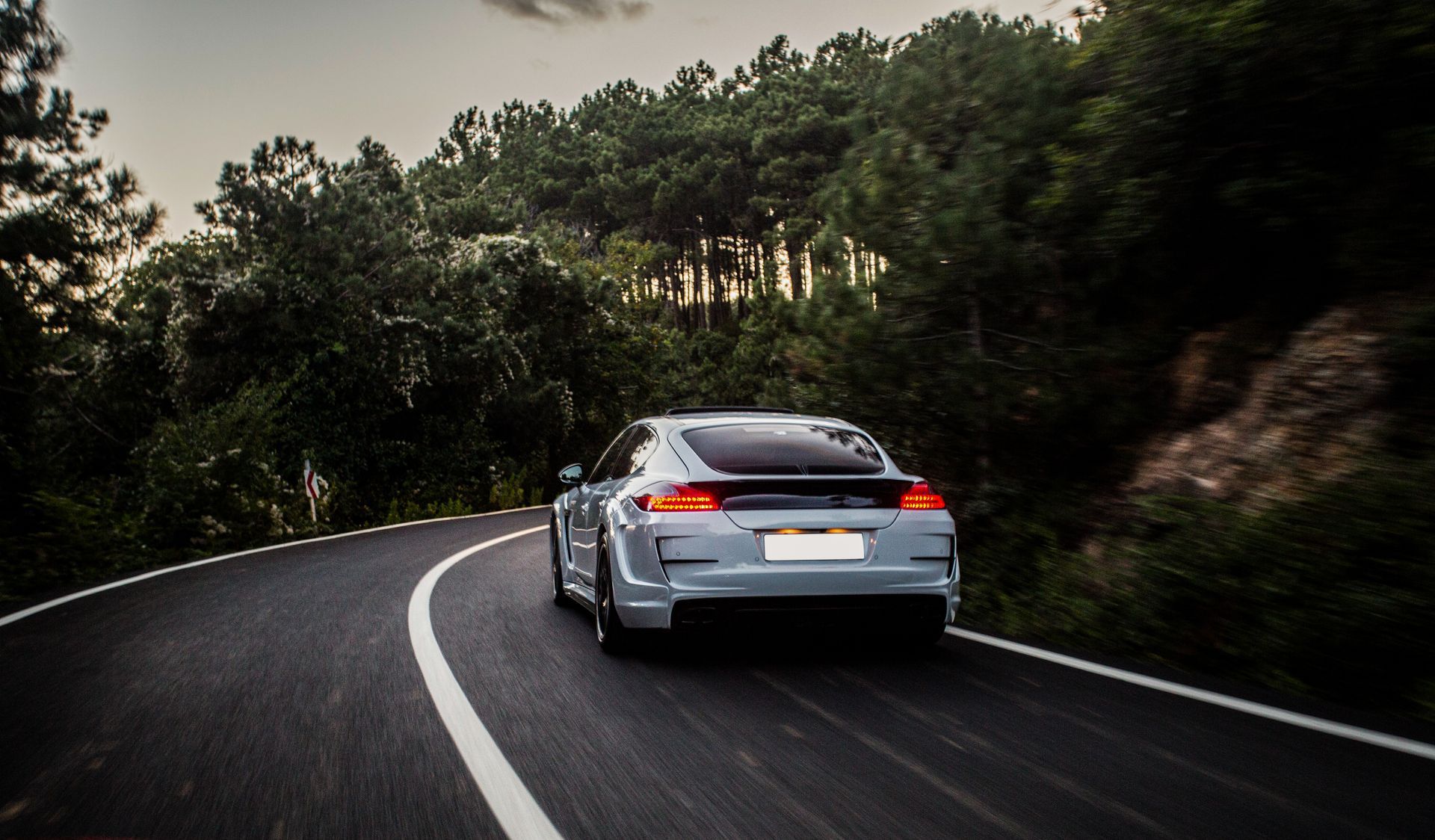 Um carro esportivo branco percorre uma estrada pavimentada sinuosa, cercada por uma exuberante floresta verde ao entardecer.