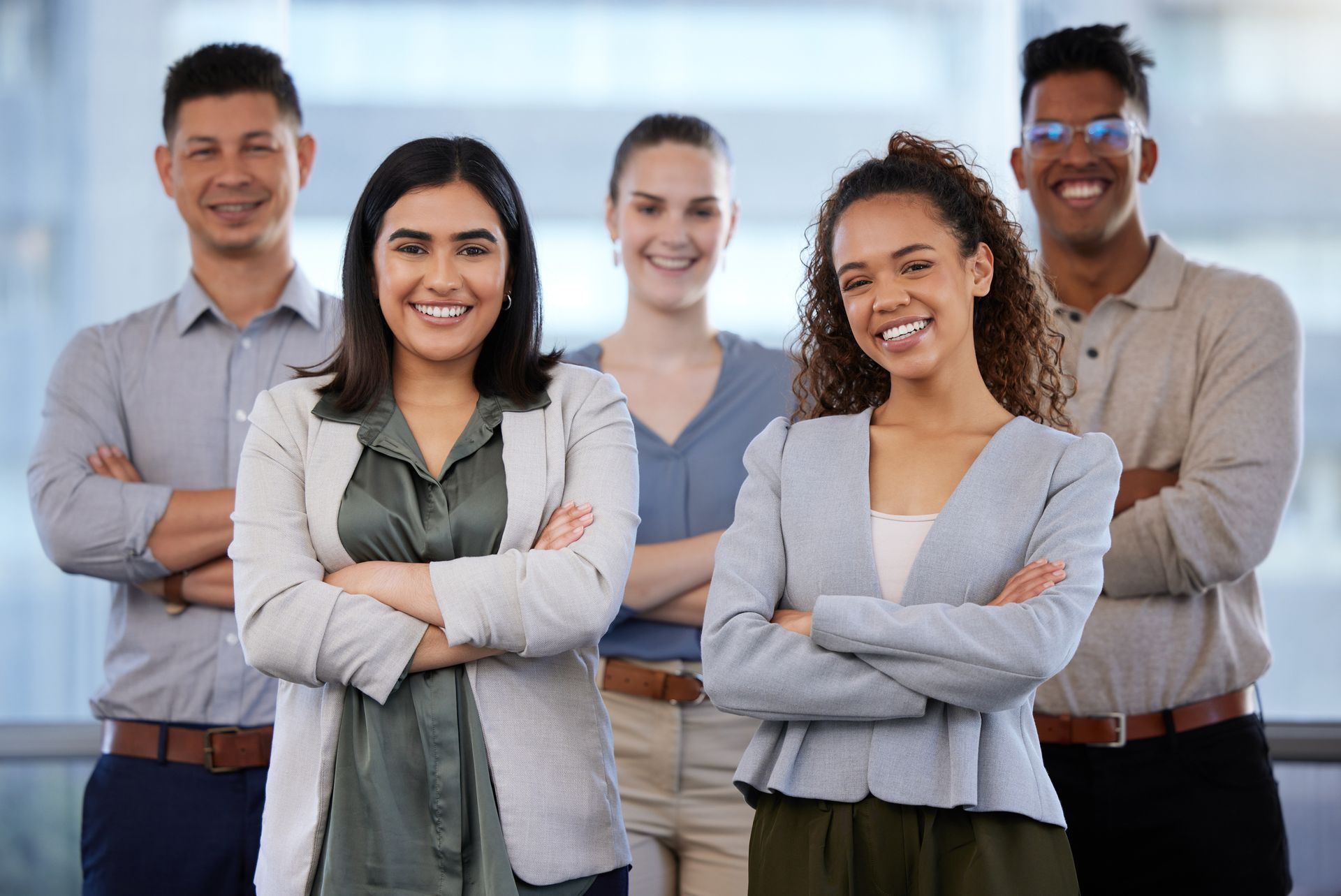 Um grupo de cinco profissionais, de braços cruzados, sorrindo para a câmera em um ambiente de escritório bem iluminado.