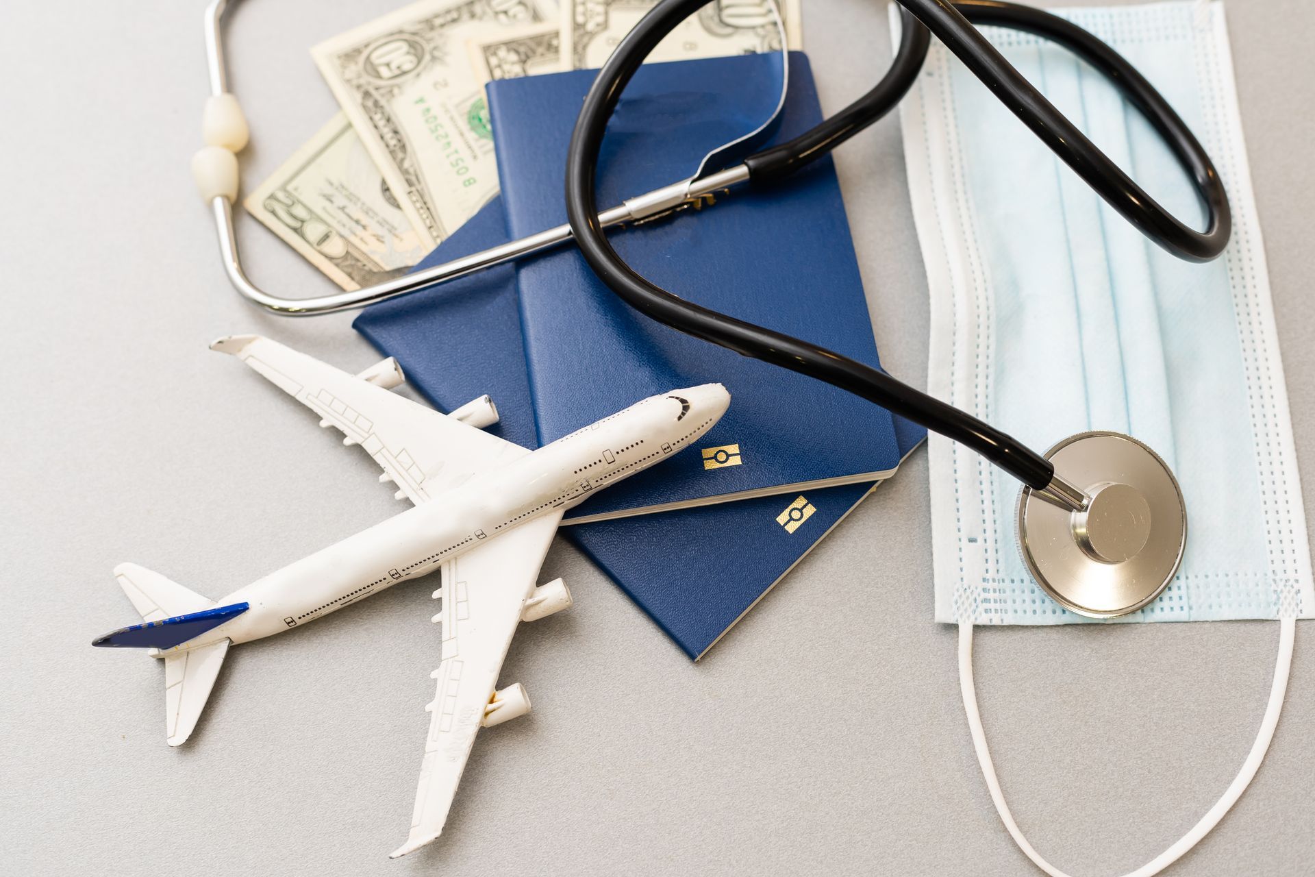 Avião de brinquedo, dois passaportes azuis, dinheiro, um estetoscópio e uma máscara facial sobre uma superfície cinza.
