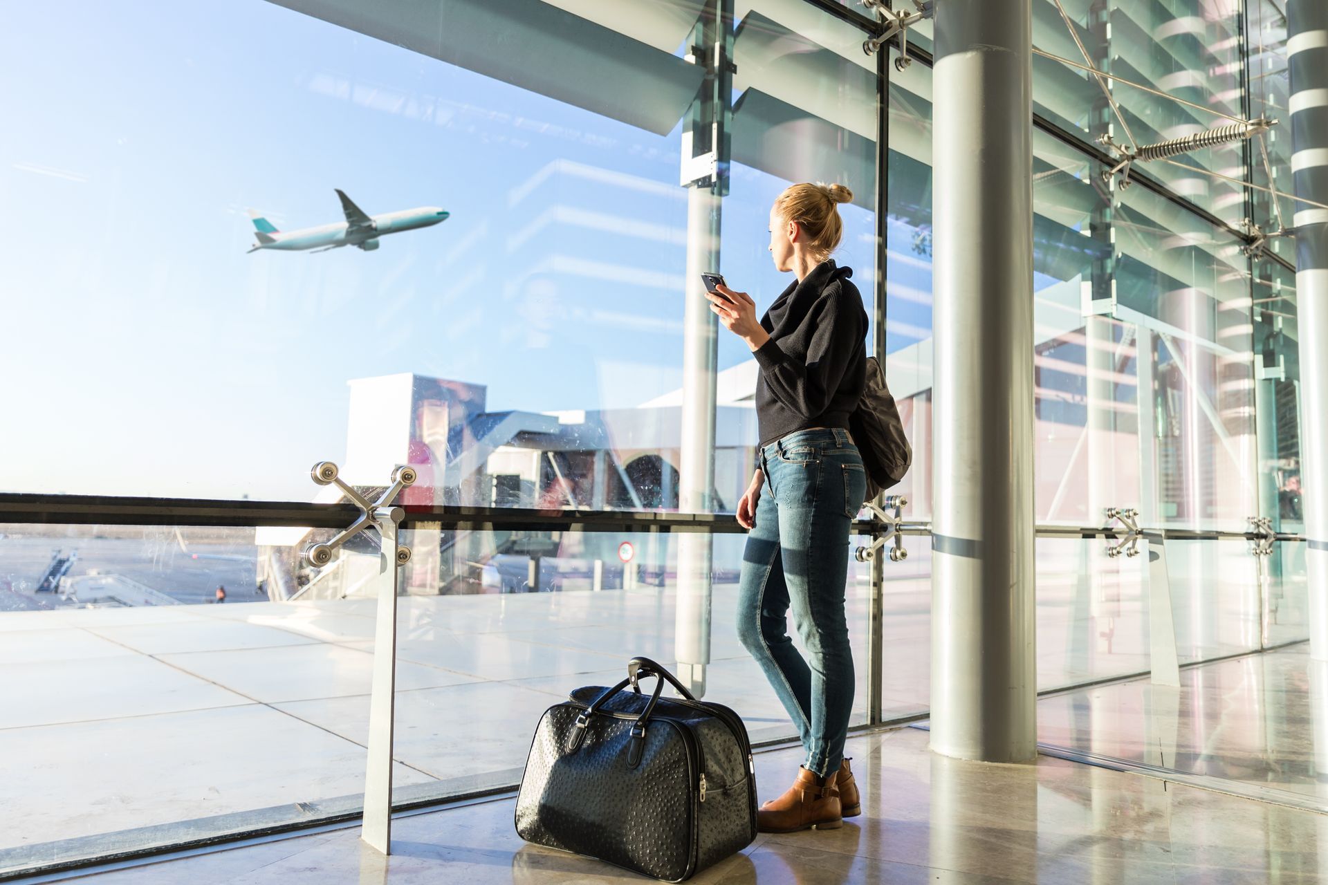 Viajante com mala de viagem está em terminal de aeroporto, olhando através de grande janela de vidro para um avião no céu.