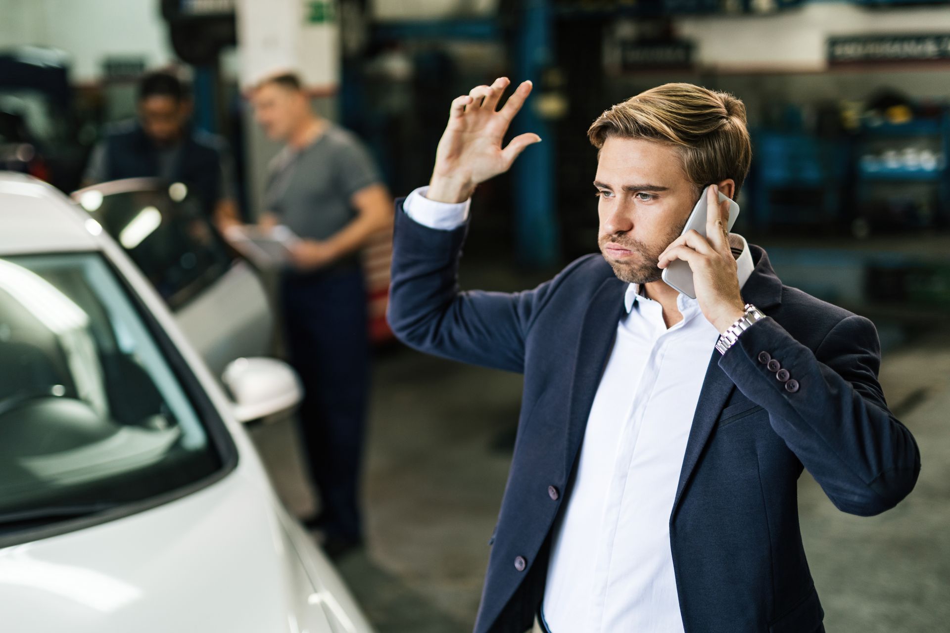 Homem de blazer azul falando ao telefone ao lado de um carro branco, com aparência estressada em uma concessionária.