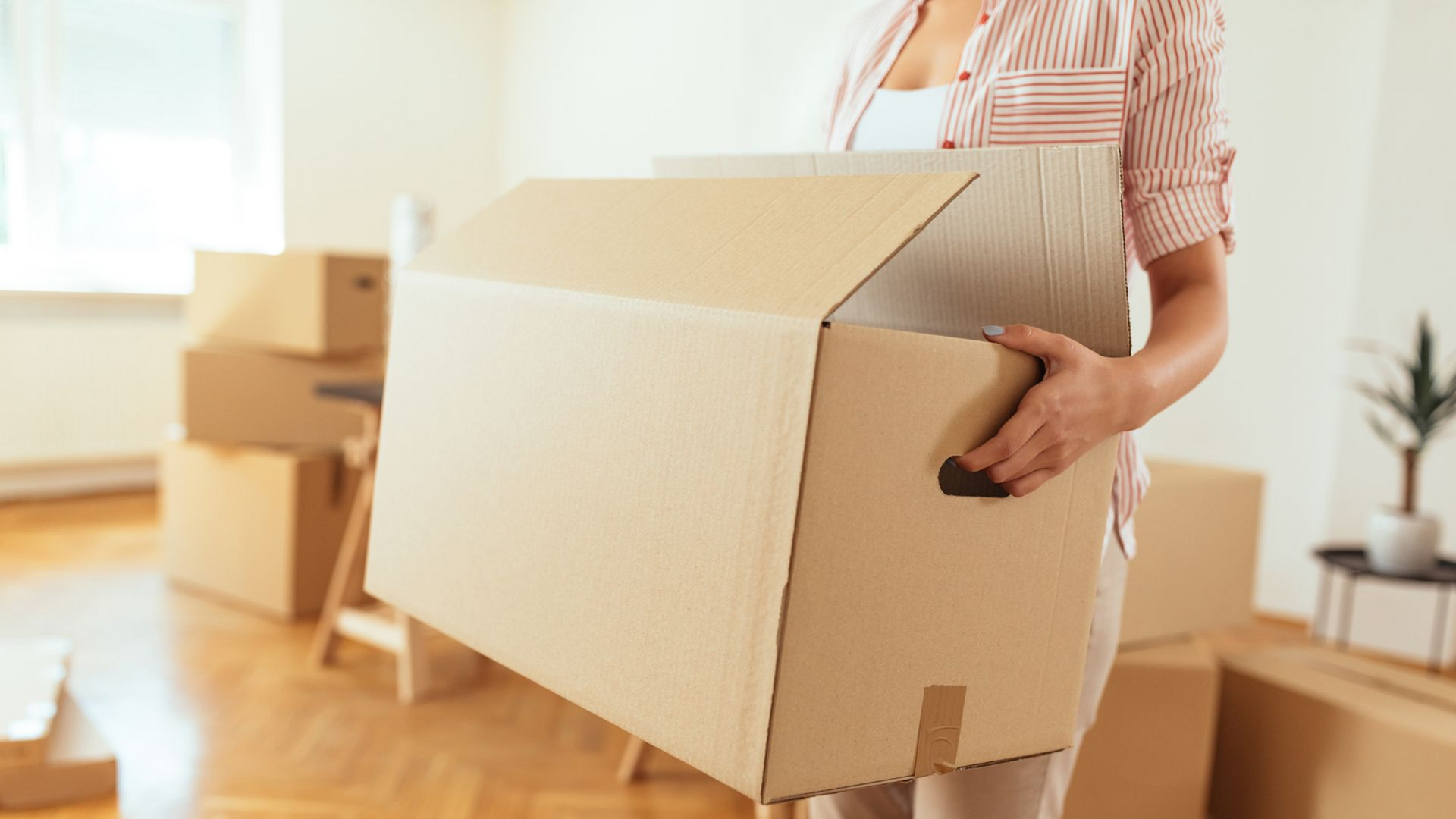 Person carrying a large cardboard moving box in a bright room with more boxes nearby
