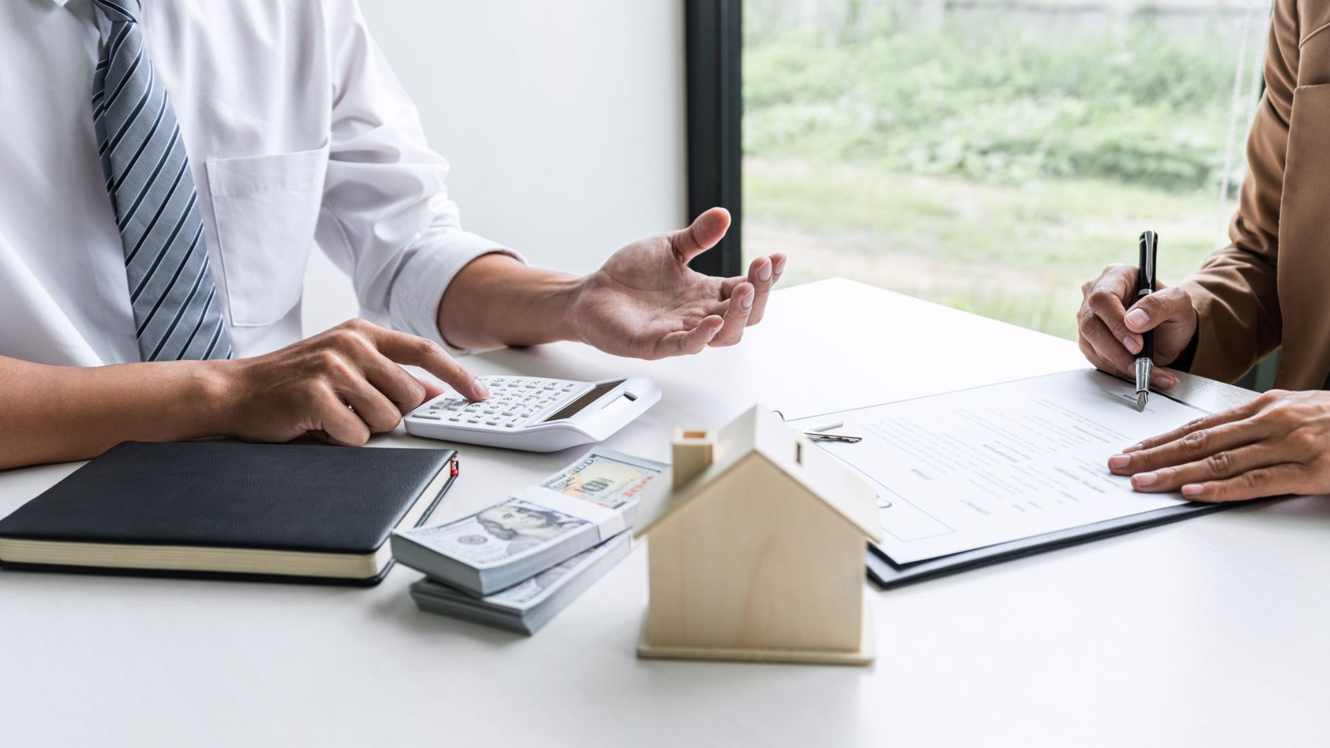 Two people reviewing financial documents at a desk with calculator, notebook, and house model