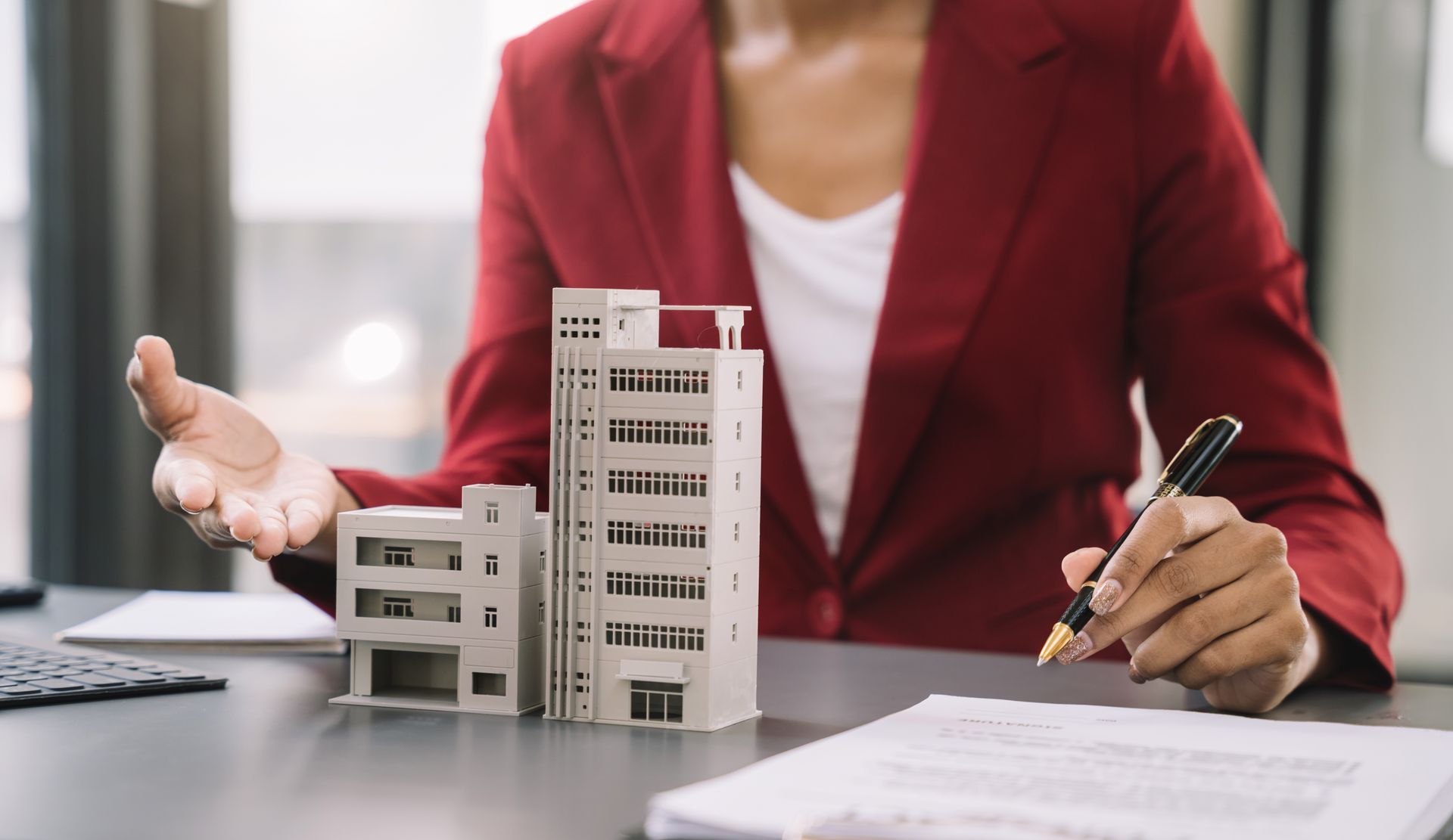 Person in red blazer presenting building models and writing on paperwork at a desk