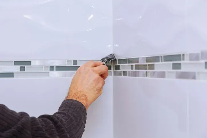 Hand placing tile on a white bathroom wall with a gray mosaic border