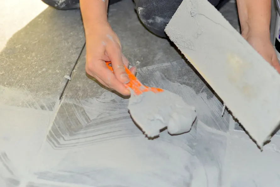 Hands using a trowel to spread gray plaster on a floor, with a board nearby.
