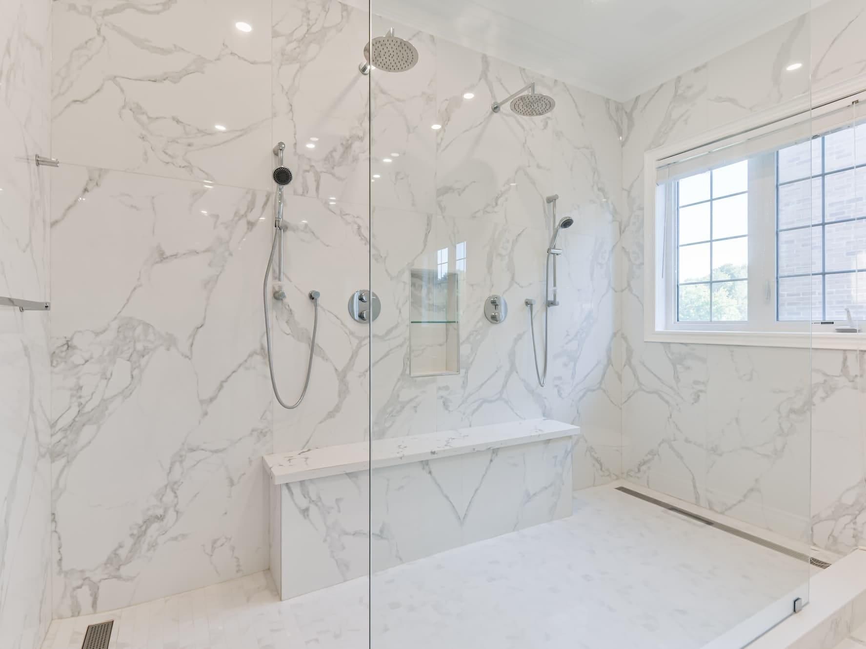 Bright white marble bathroom with glass shower, built-in tub, and window with shutters.