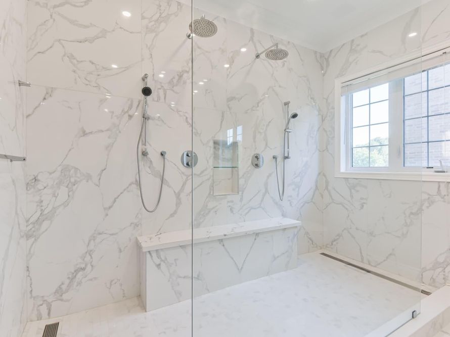 Bright white marble bathroom with glass shower, built-in tub, and window with shutters.