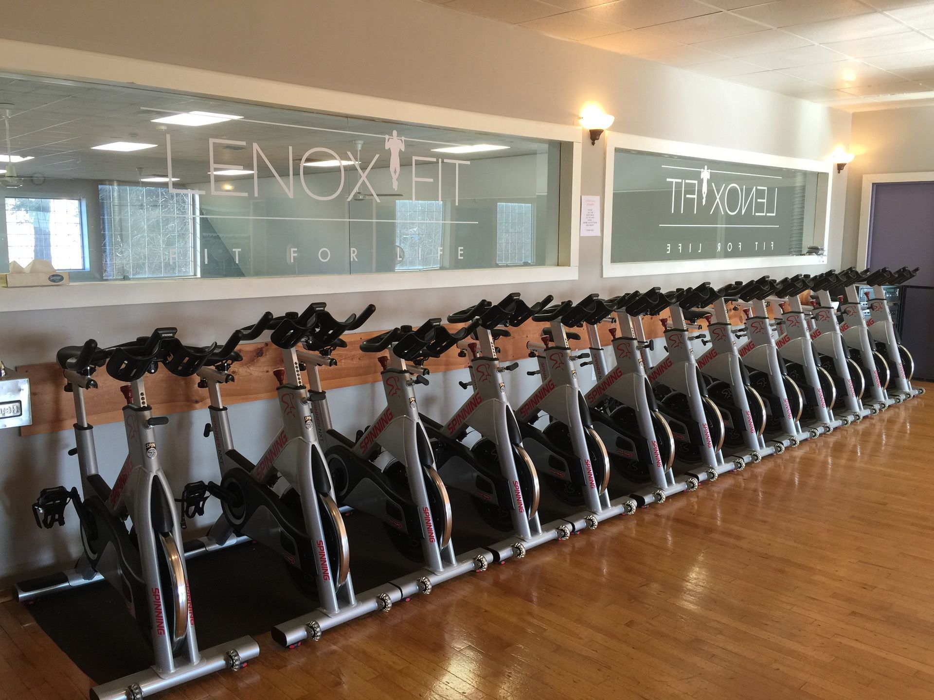 A row of stationary exercise bikes lined up in a studio with mirrored walls featuring the Lenox Fit logo.