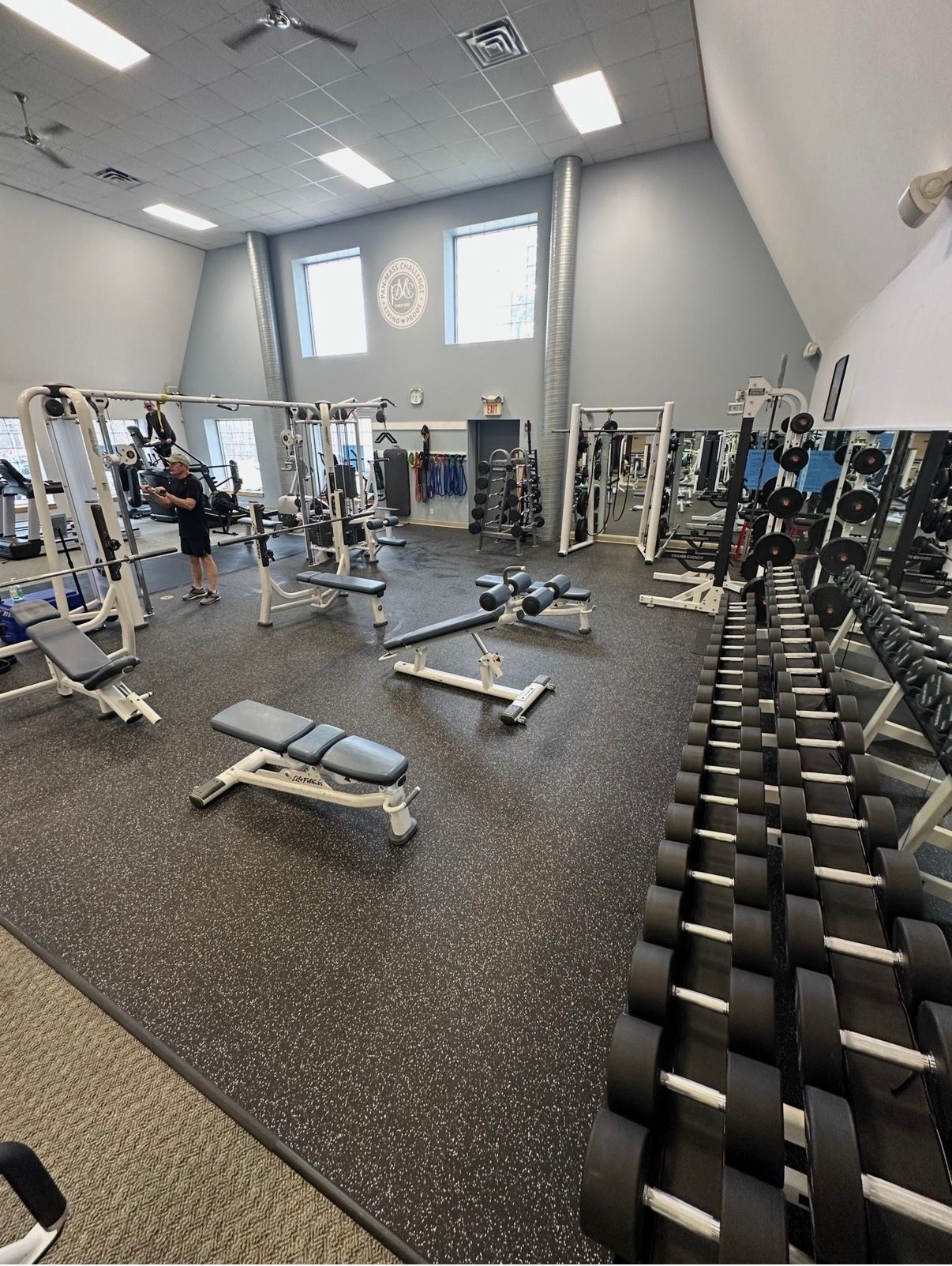 Gym interior with rows of dumbbells on a rack, workout benches, and weight machines under high ceilings and windows.