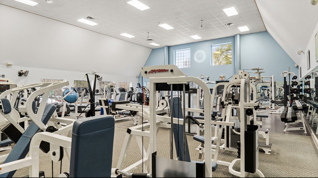A gym filled with white strength training machines under a high, sloped ceiling with bright overhead lighting.