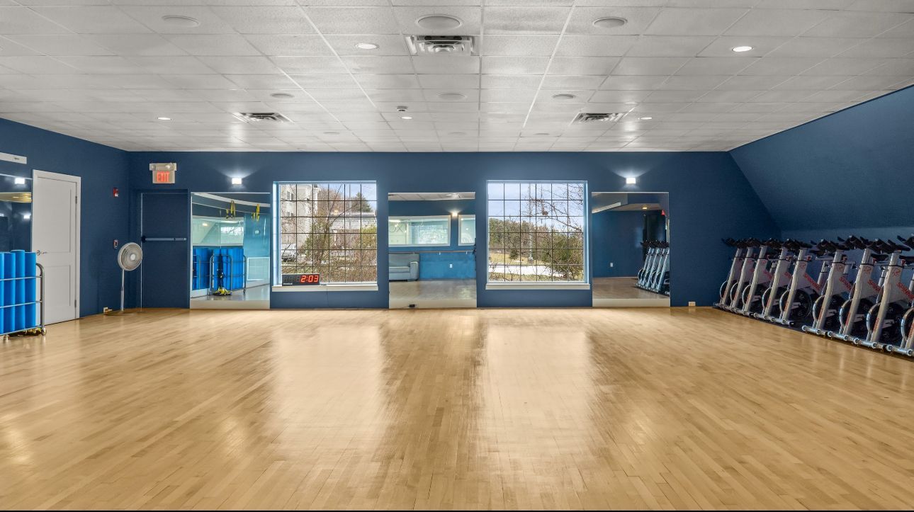 A spacious fitness studio with blue walls, light wood floors, windows, and a row of stationary bikes along the right wall.