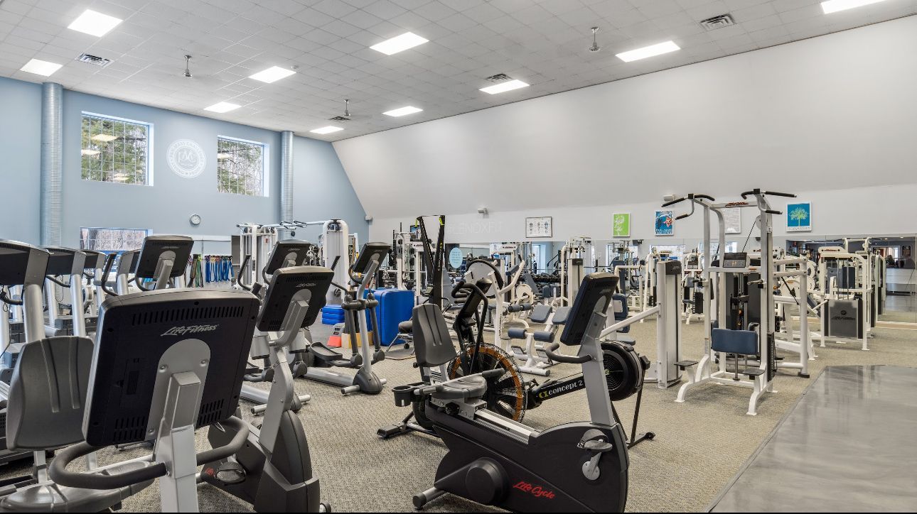 A spacious, brightly lit gym filled with various weight training and cardio exercise machines arranged on a gray floor.
