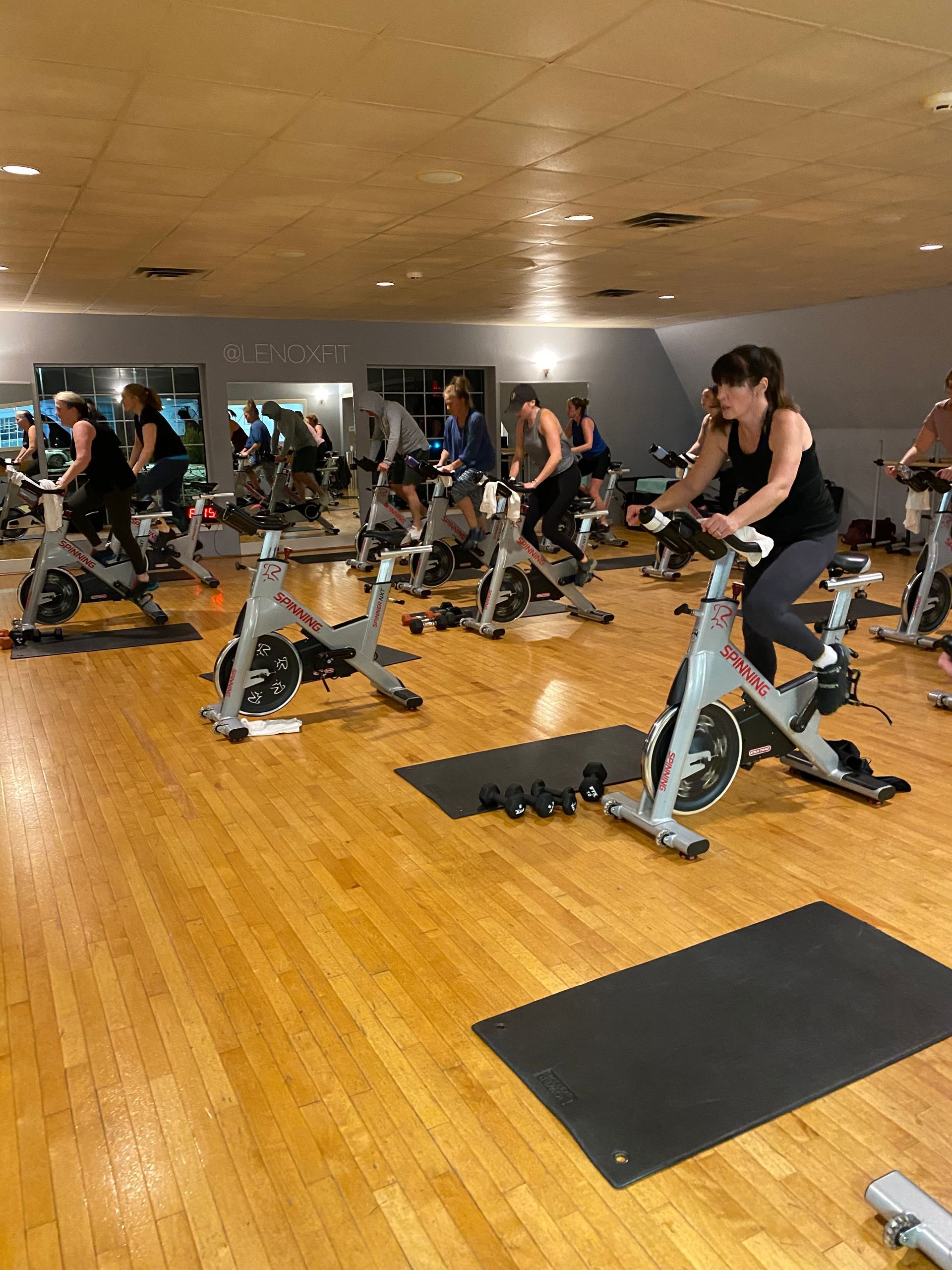 A group of people attending an indoor cycling class in a gym with hardwood floors and mirrors.