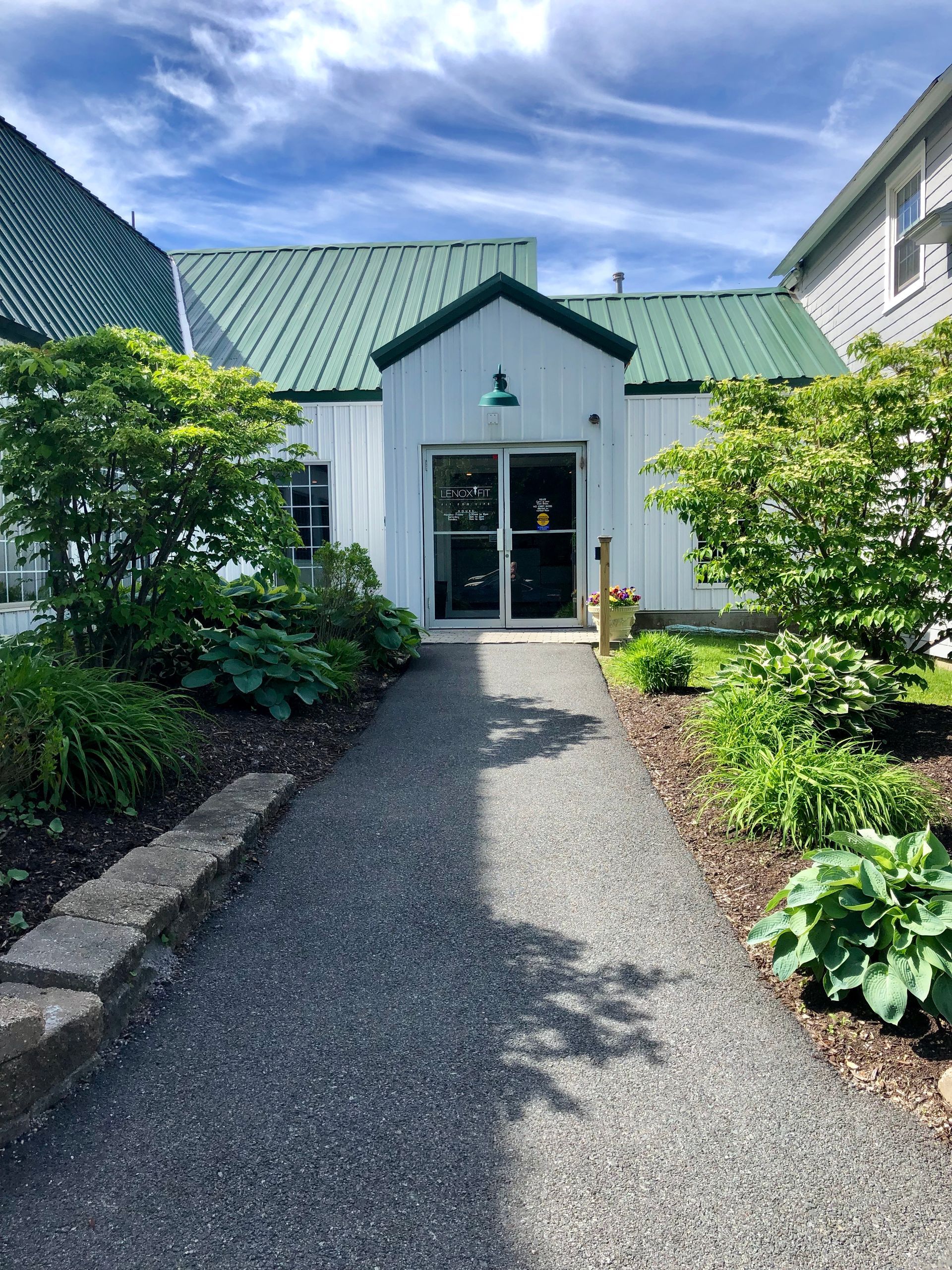 A gravel path leads to the white, green-roofed entrance of a building surrounded by lush green bushes and trees.