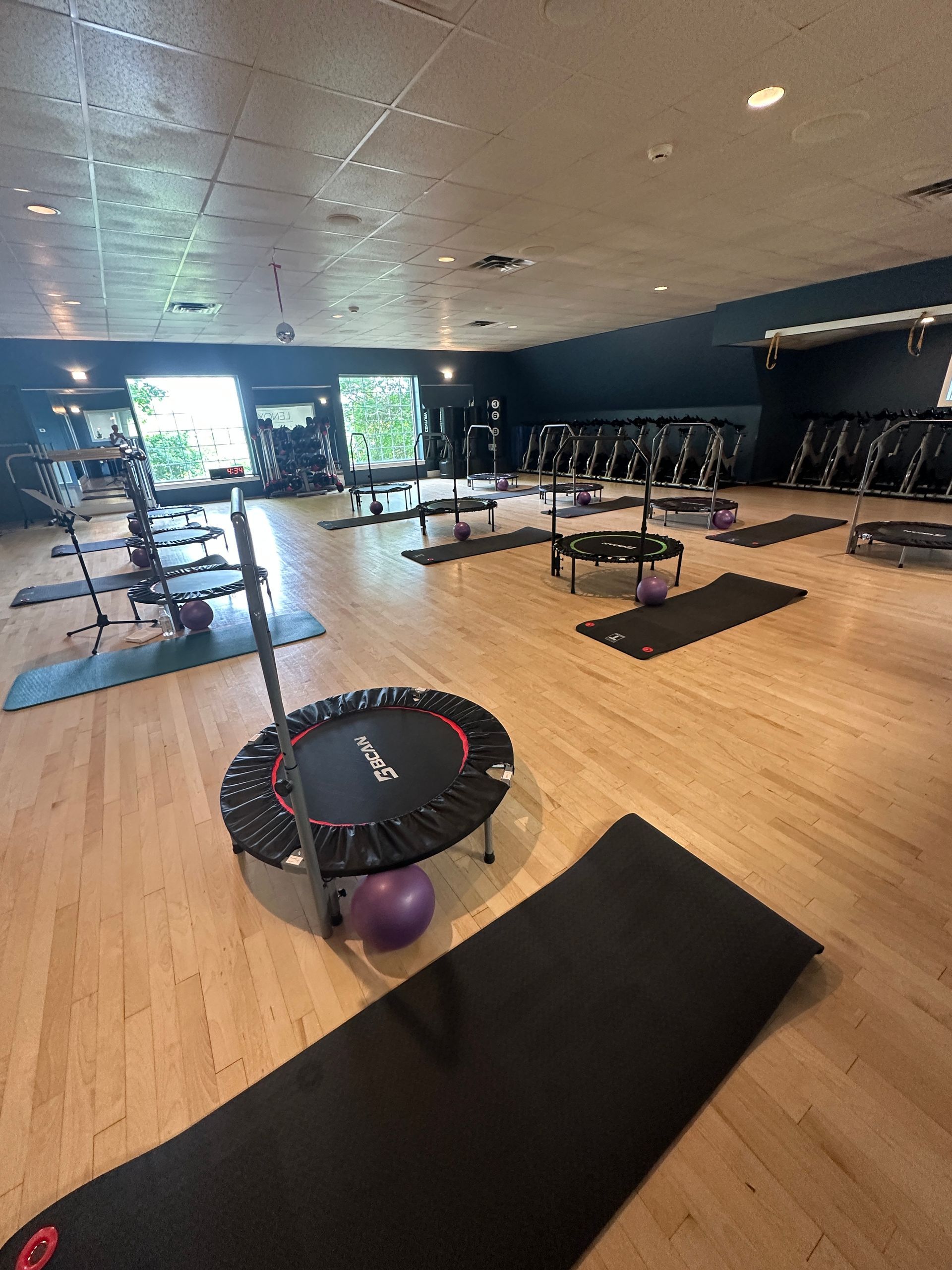 A fitness studio interior with rows of small trampolines, exercise mats, and purple medicine balls on a light wood floor.