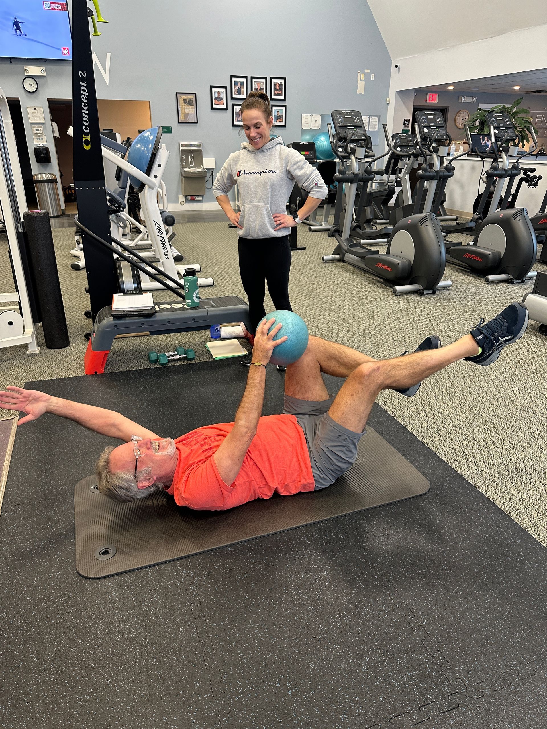 A person performs a core exercise with a blue ball on a gym mat while a trainer observes nearby.