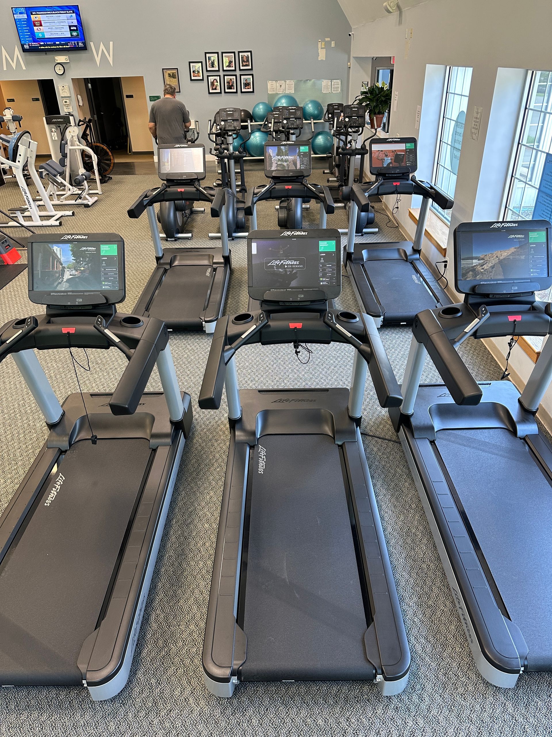 A row of three identical black treadmills with digital screens, situated in a gym with stationary bikes in the background.