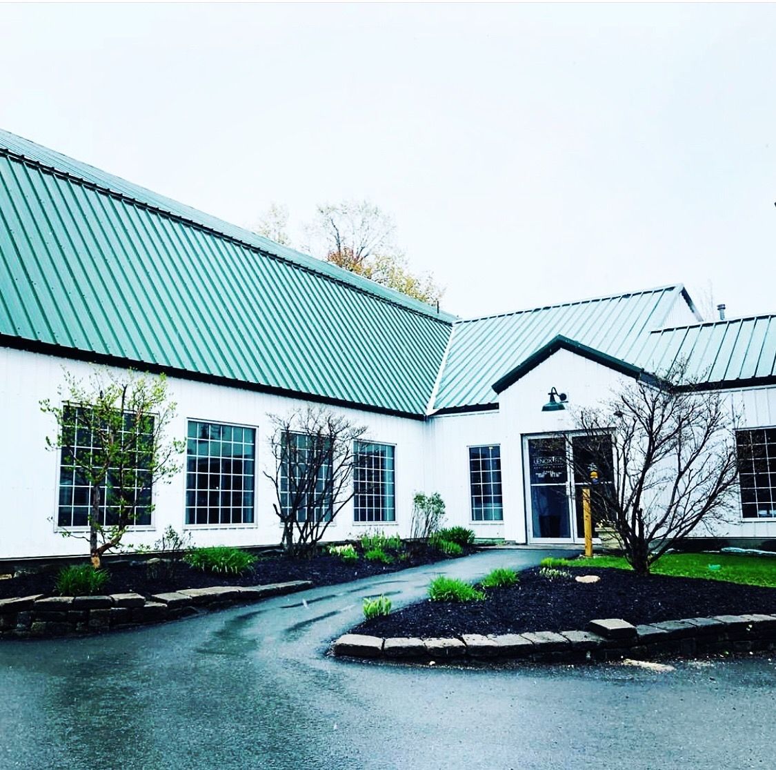 A one-story white building with a green metal roof, multiple windows, and a paved walkway in front of the entrance.