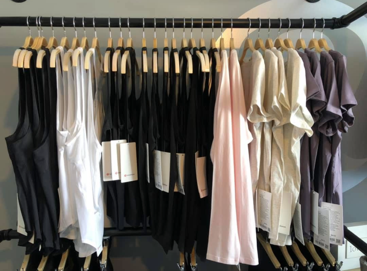 A retail clothing rack displays various tank tops and t-shirts in black, white, light pink, and mauve colors.