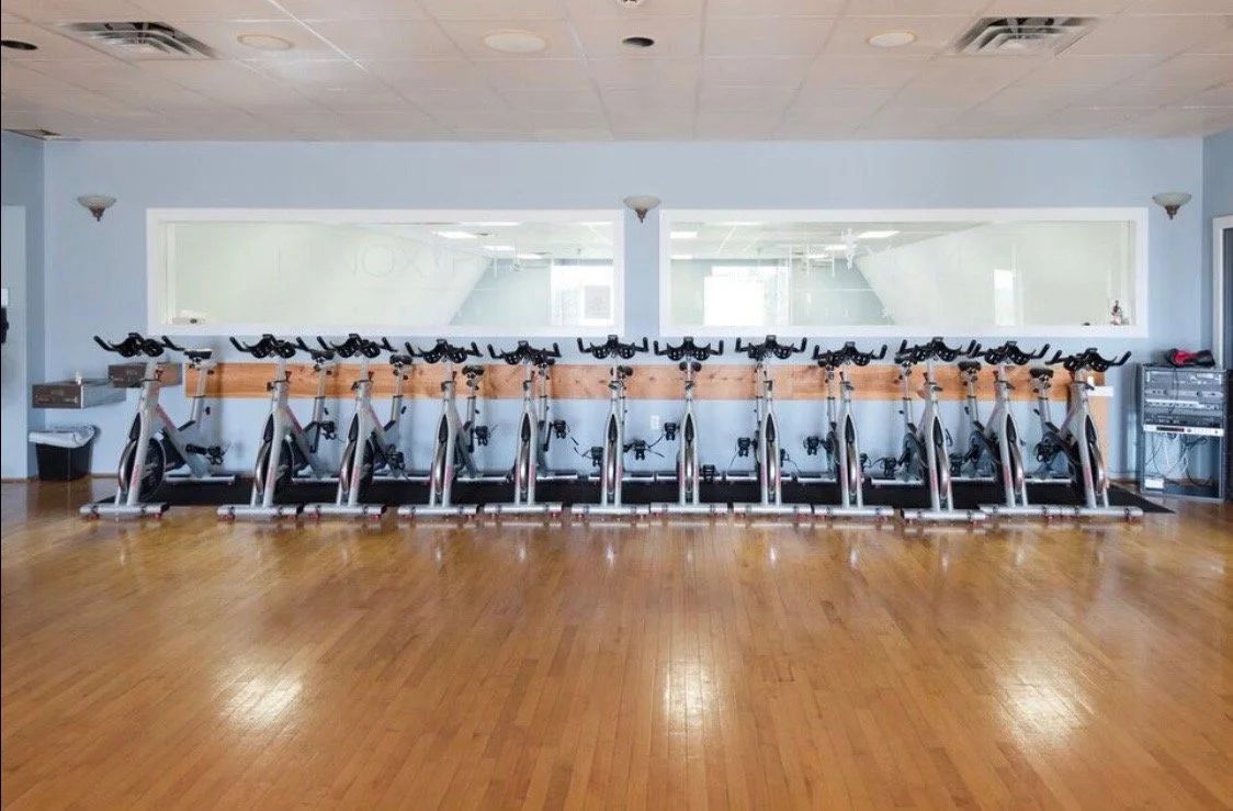 A row of stationary exercise bikes lined up on a hardwood floor in a fitness studio with mirrors on the back wall.