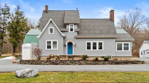 A two-story grey house with a blue door, a shingled roof, two chimneys, and a stone retaining wall in front.