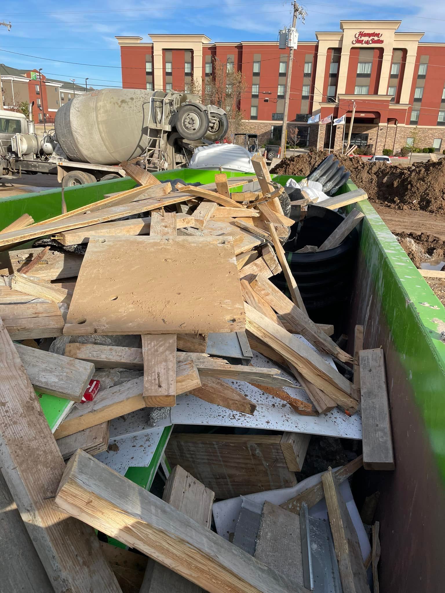 Construction debris including wood scraps and plastic pipes fill a large, bright green dumpster in front of a brick hotel.