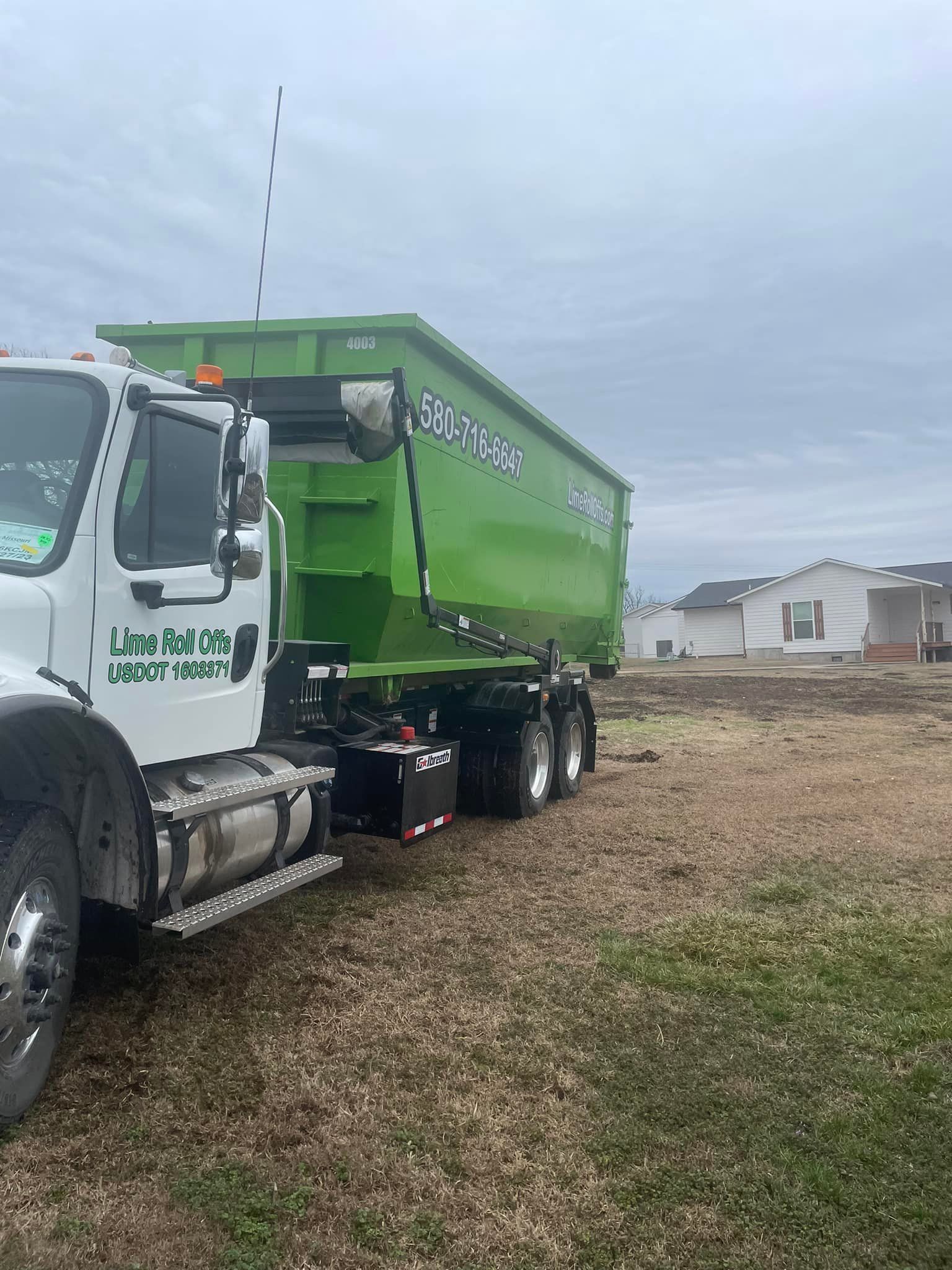 A white roll-off truck with a large lime green dumpster container parked at a residential construction site.
