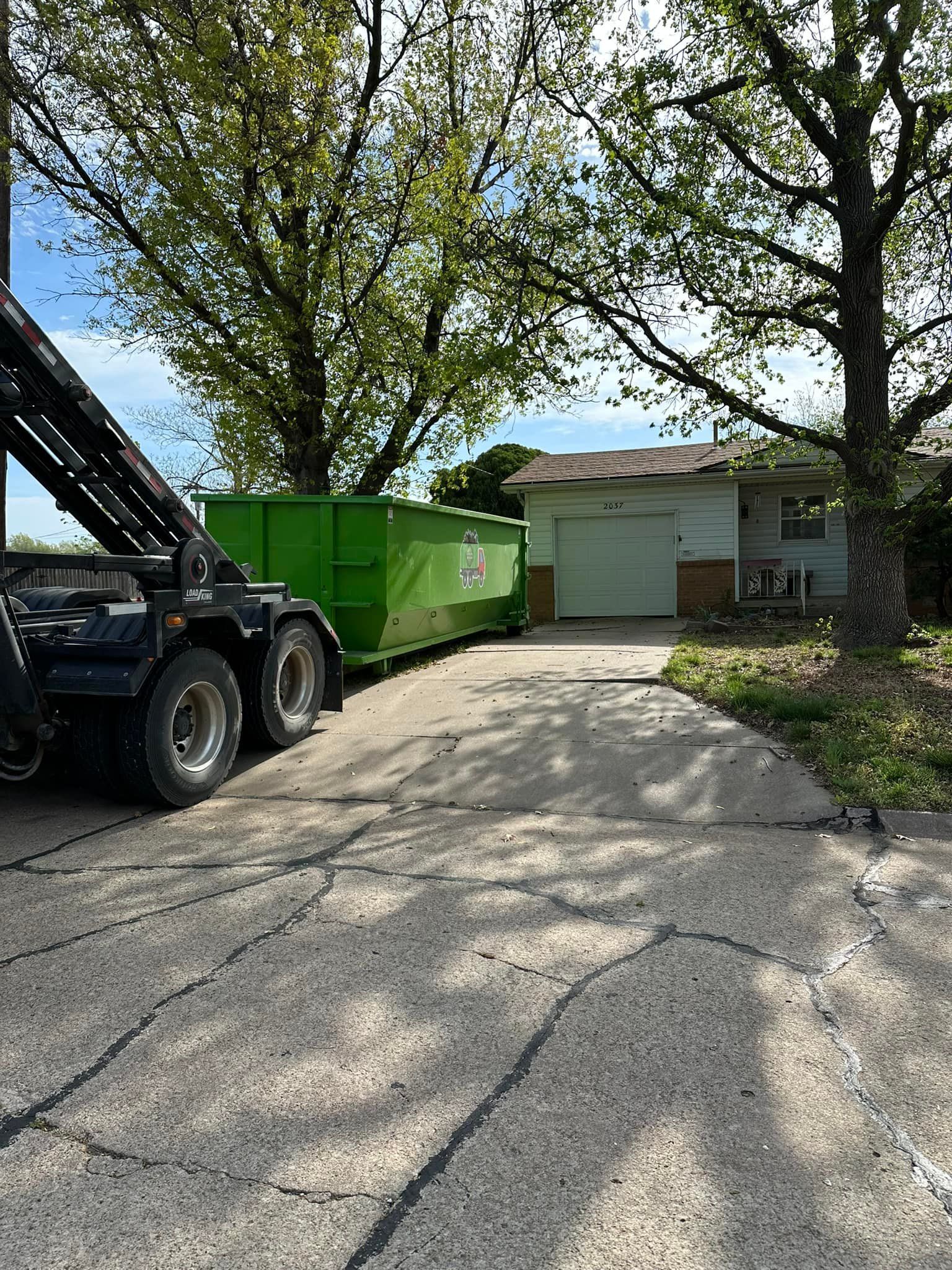 A bright green dumpster is positioned on a residential driveway in front of a house, being dropped off by a service truck.