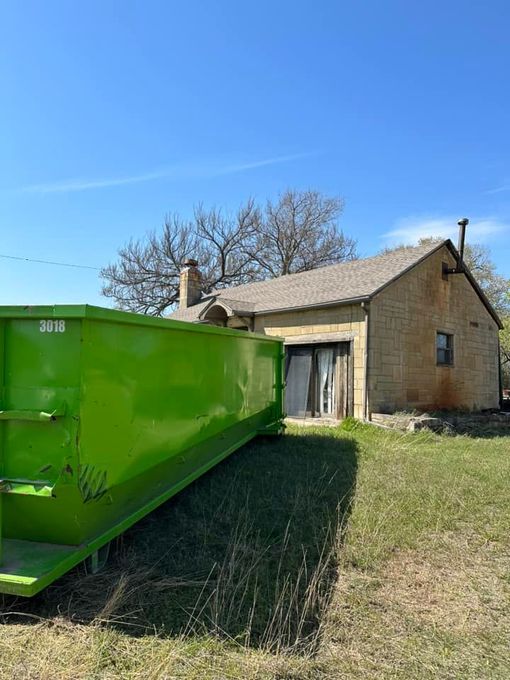 A bright green dumpster sits on a grassy field in front of a rustic, light-brown stone building under a clear blue sky.