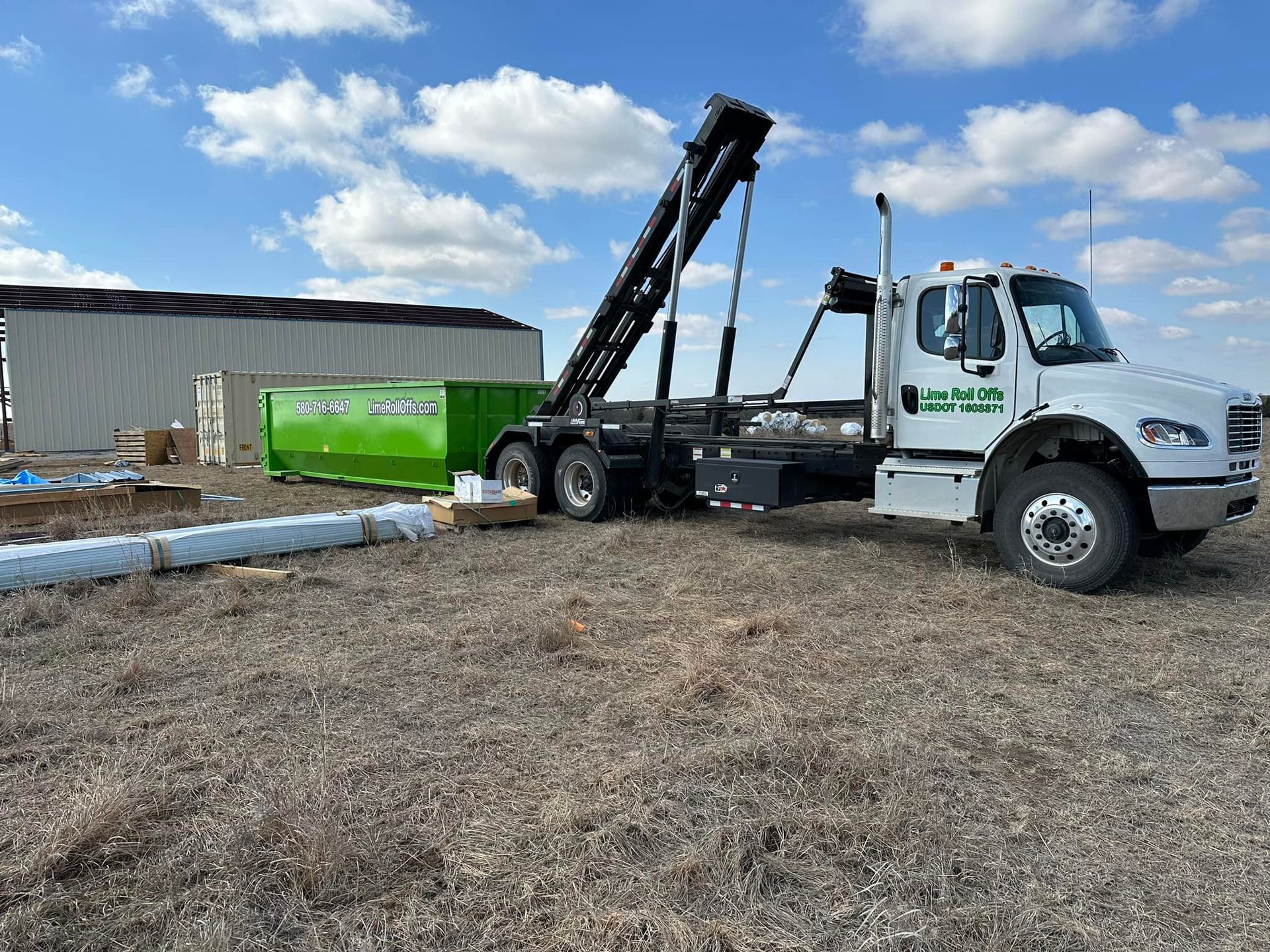 A white roll-off truck is lowering a bright green dumpster onto a dirt field in front of a metal building.