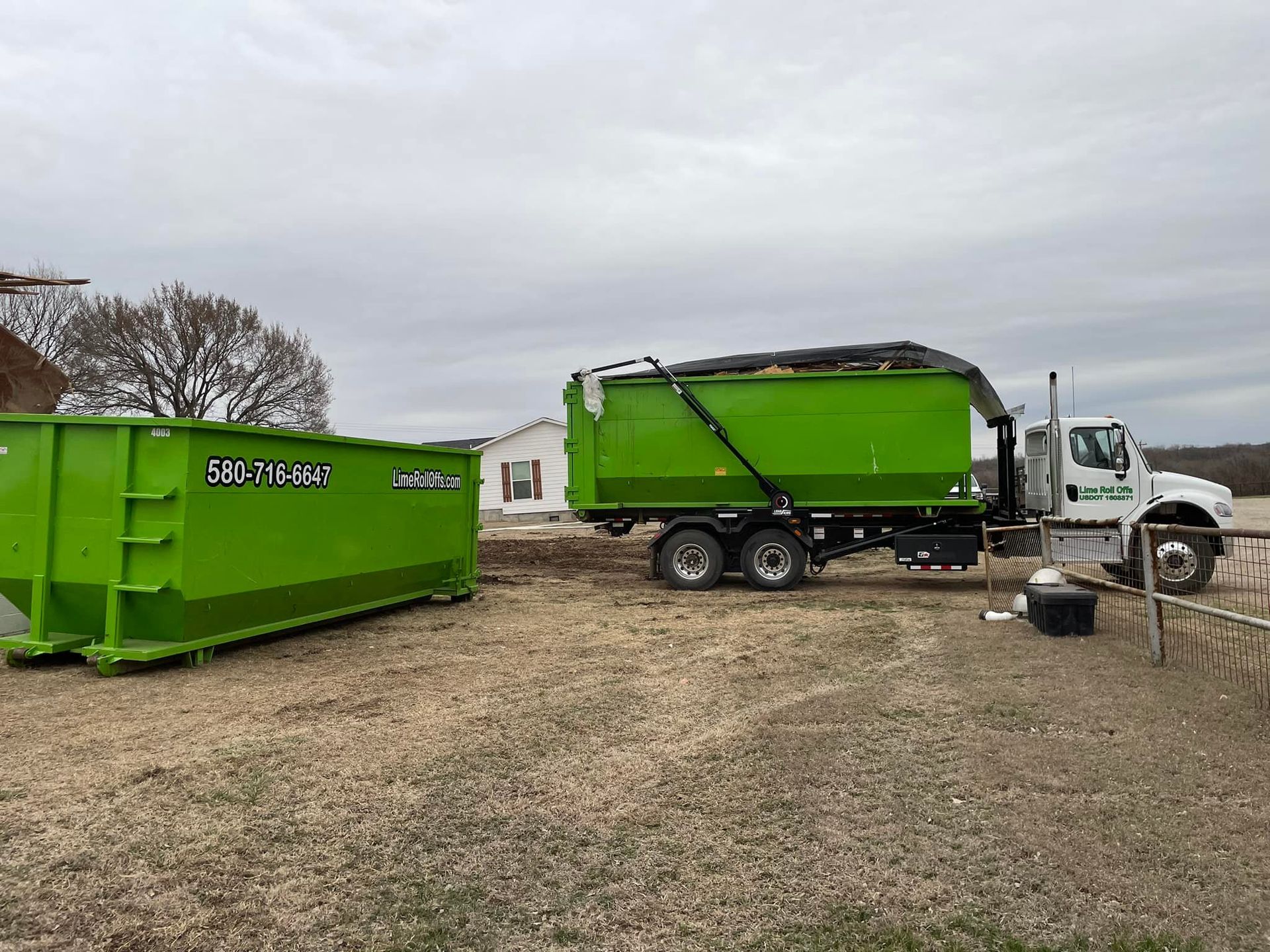 A white truck hauls a bright green dumpster, with a second matching green dumpster parked on a dirt lot near a house.