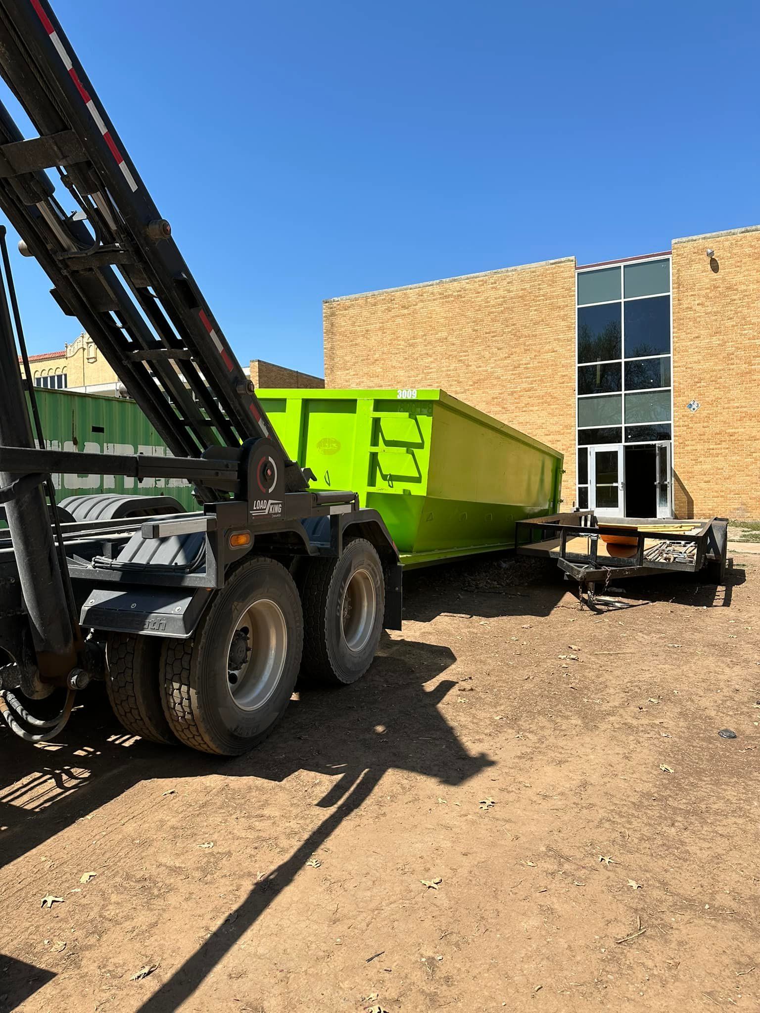 A bright green dumpster being positioned by a roll-off truck in front of a brick building on a dirt lot.