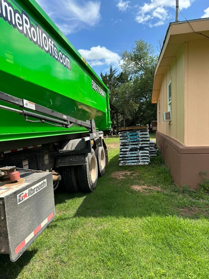 A bright green roll-off dumpster truck parked on a lawn next to a building, with a stack of wooden pallets nearby.