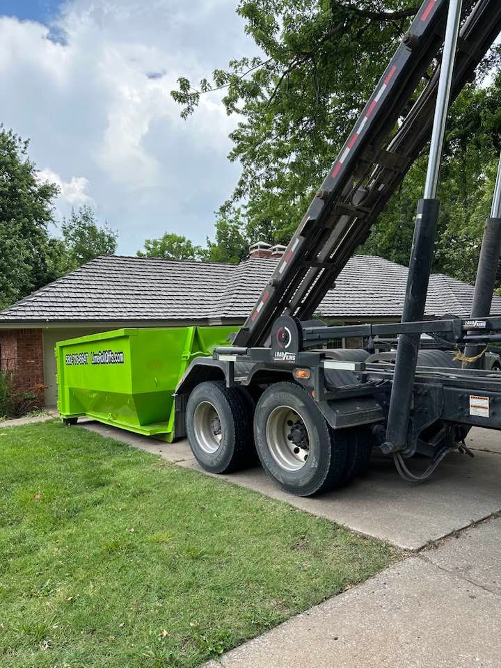 A bright green dumpster being lowered from a truck onto a concrete driveway in front of a residential home.