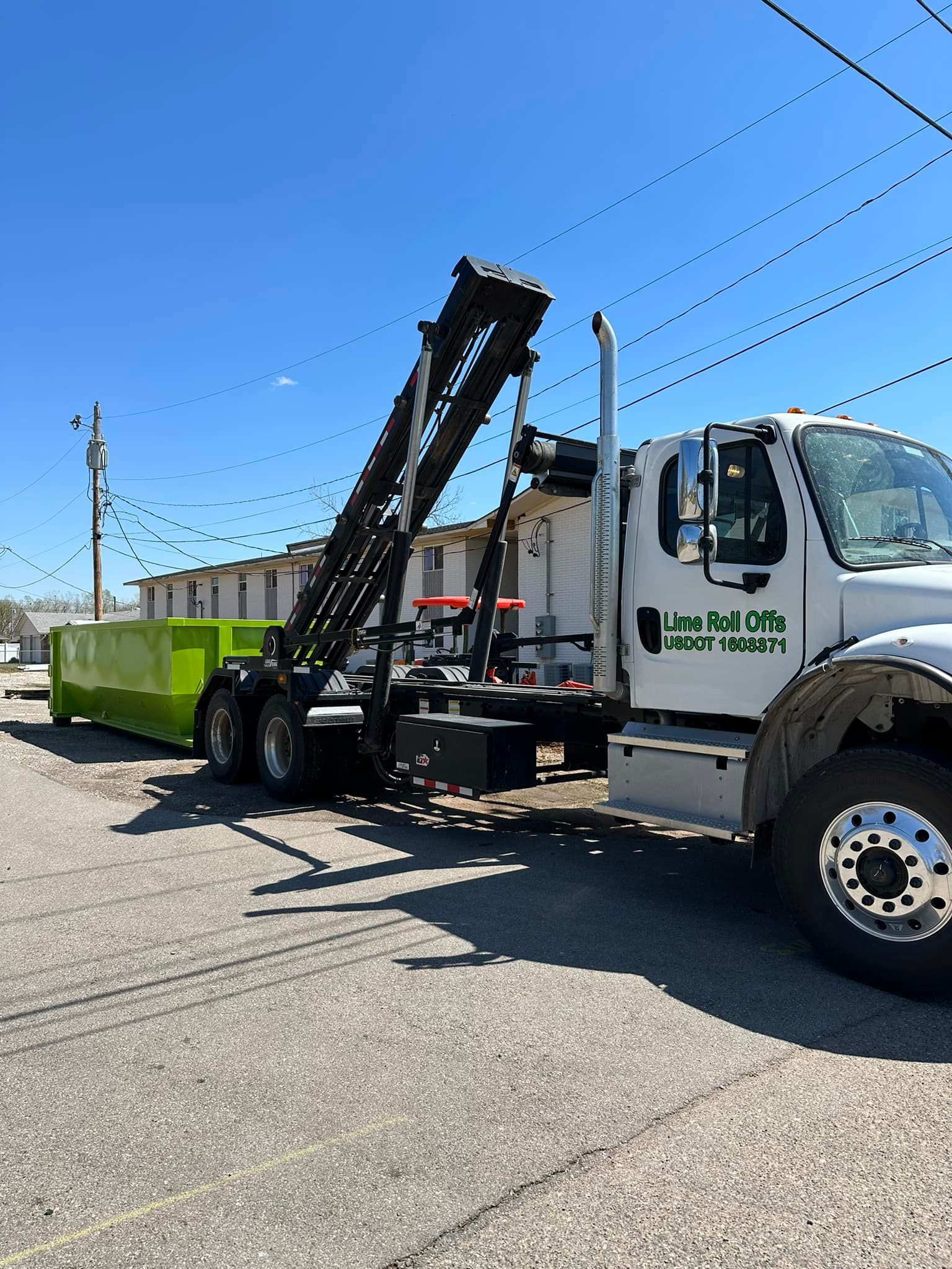 A white roll-off truck hauling a bright green dumpster in a sunny parking lot.