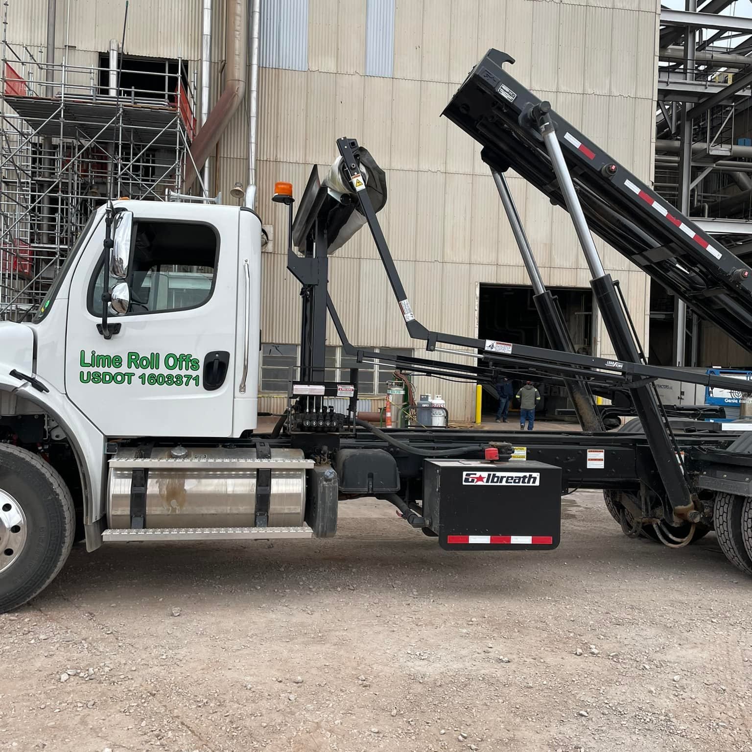 A white roll-off dump truck with its hydraulic lifting mechanism raised, parked on a gravel lot near an industrial site.
