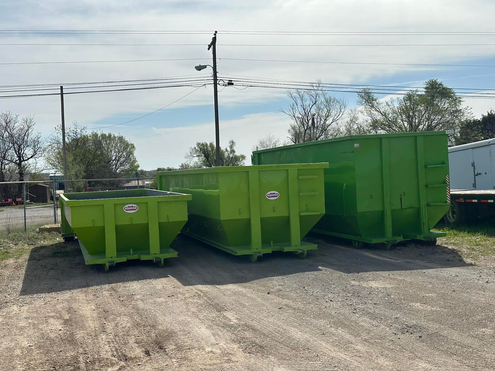 Three lime-green roll-off dumpsters of varying sizes lined up on a gravel lot under a clear sky.