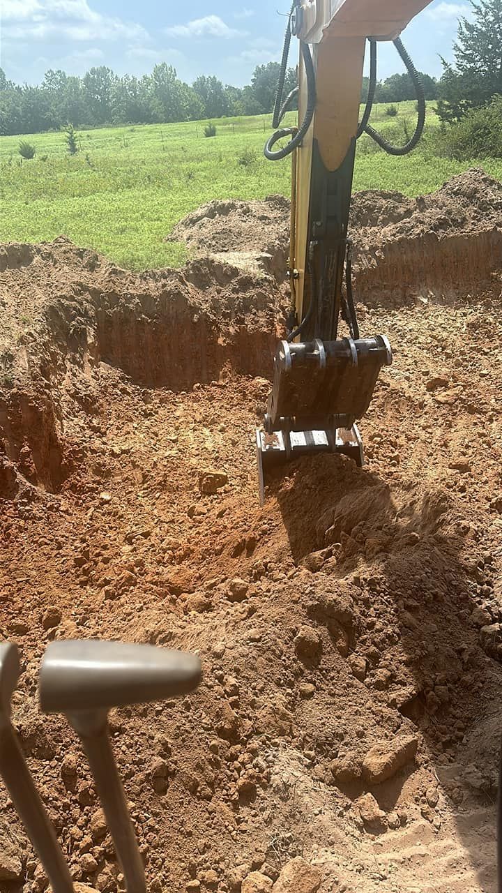 An excavator bucket digs into a mound of brown soil in an open field under a blue sky.