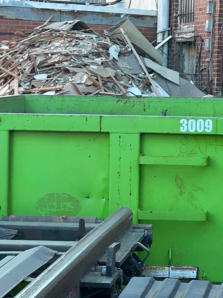 A large green construction dumpster sitting in front of a pile of building debris near a brick wall.