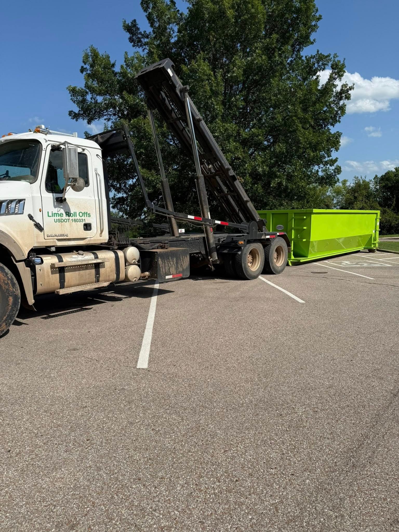 A white roll-off truck in a gravel parking lot loading a bright lime-green dumpster onto its flatbed.