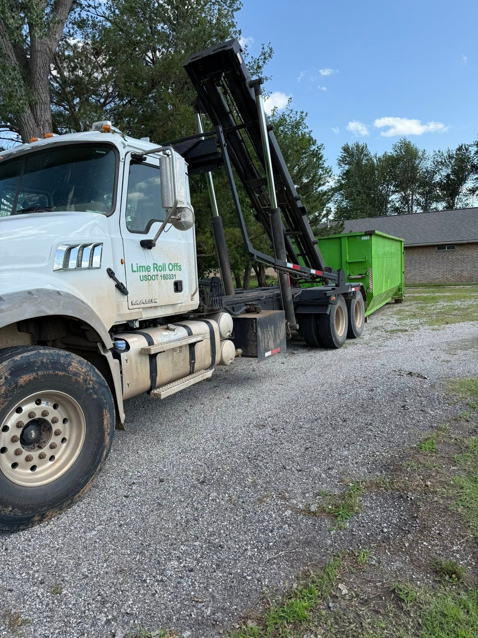 A white roll-off dump truck with a mechanical arm loading a bright green dumpster onto its bed on a gravel lot.