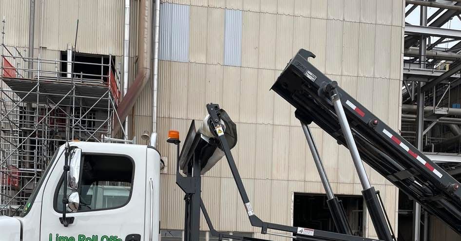 A white roll-off truck with its hydraulic arm raised at a construction site with scaffolding against a building.